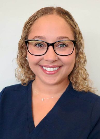 A smiling person with curly hair and glasses, wearing a navy blue scrub top against a neutral background.