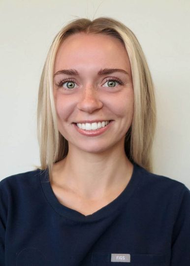 A person with blonde hair and a friendly smile, wearing a dark blue scrub top against a plain light background.