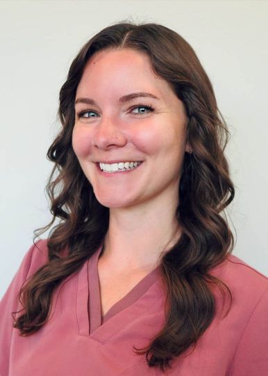 A person with long, dark, wavy hair, wearing a dusty rose-colored v-neck scrub top, smiles against a plain white background.