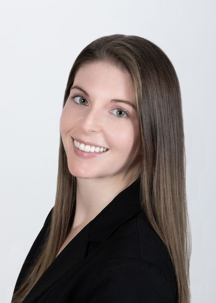 Headshot of a person with long brown hair, wearing a black blazer, smiling against a plain white background.