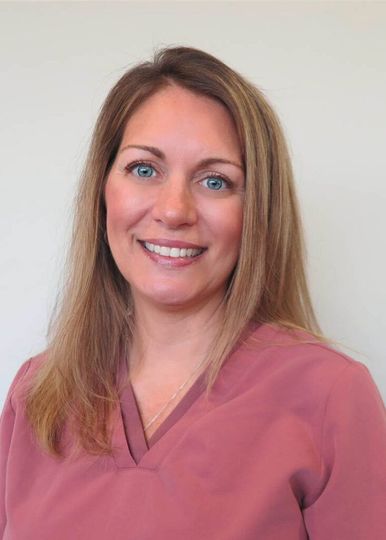A smiling professional with long brown hair wearing a pink V-neck scrub top against a plain light gray background.