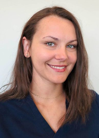 A smiling person with long brown hair, wearing a dark blue scrub top, posing against a plain light-colored background.