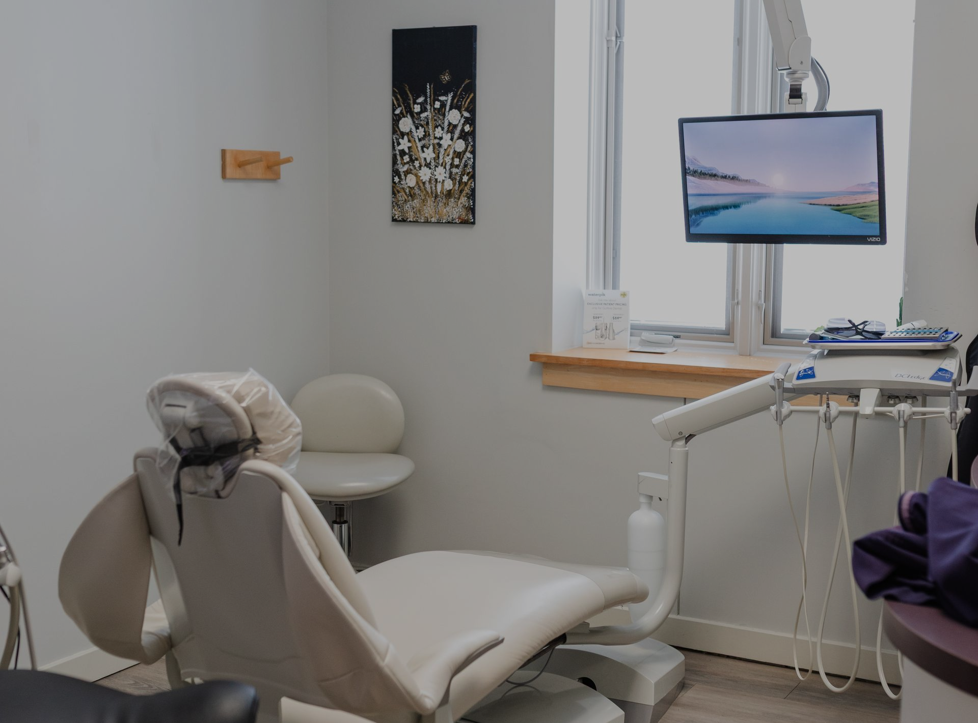 A dental examination room with a patient chair, dental equipment, a monitor displaying a landscape, and wall art.