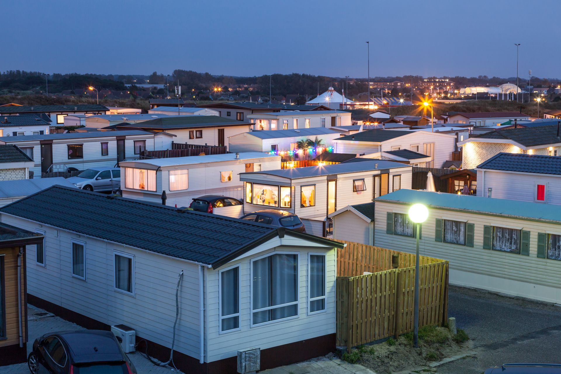 Mobile home community at dusk with lit windows, parked cars, and streetlights across the neighborhood. Mobile home community at dusk with lit windows, parked cars, and streetlights across the neighborhood.