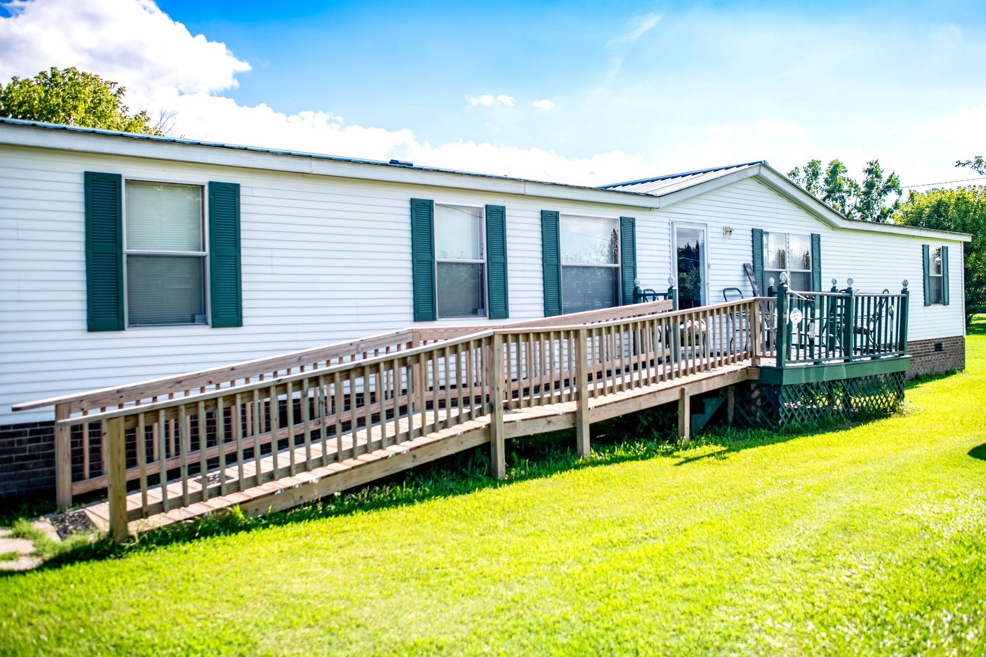 Accessible mobile home with ramp, porch, and trimmed lawn in sunny, peaceful neighborhood park.