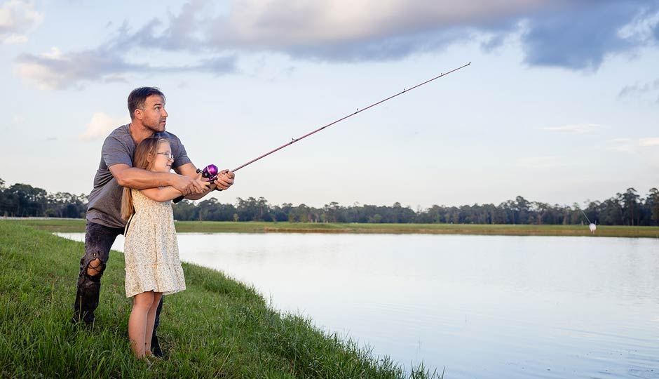 Two people fishing together on a grassy riverbank at sunset, with a calm lake and open sky.