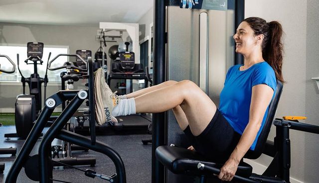 Woman exercising on a leg press machine in a gym, wearing a blue shirt and black leggings.