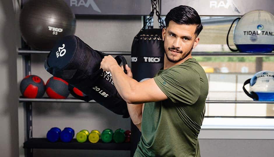 Man posing with boxing gloves in a gym, surrounded by medicine balls and fitness equipment.