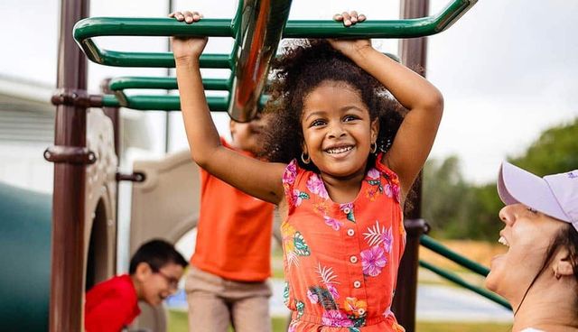Smiling child hanging from green playground bars with adults nearby