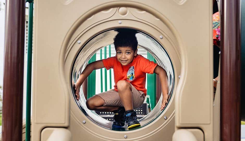 Child crouching in a playground tunnel, wearing an orange shirt and blue shorts, looking through the opening.