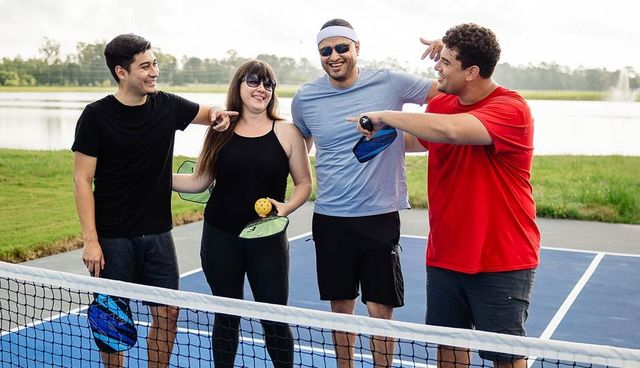 Four friends stand by a tennis net on an outdoor court, smiling and chatting with rackets in hand.