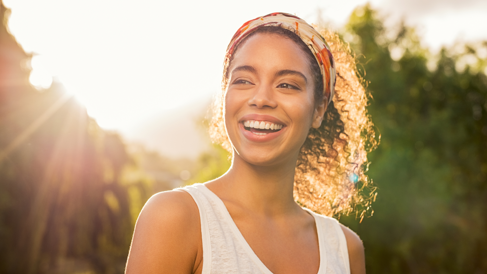 Smiling person outdoors in warm sunlight, with trees and a bright sky in the background