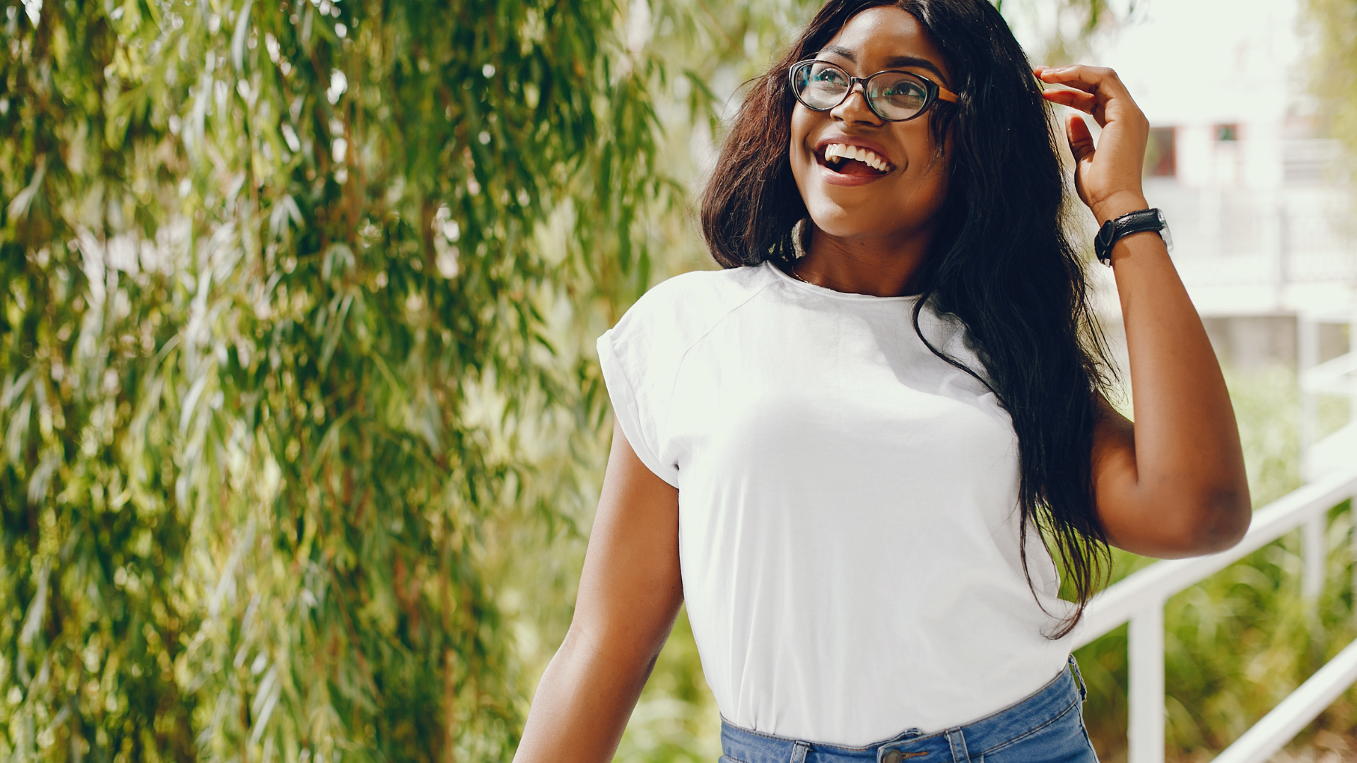 Smiling woman in a white shirt outdoors, adjusting her hair near greenery.