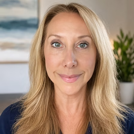 Portrait of a smiling blonde person in a navy top, standing indoors with a soft blurred background.