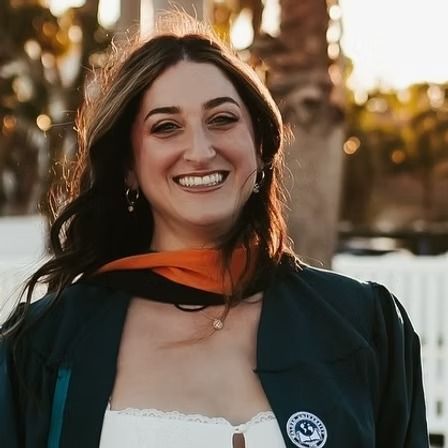 Smiling graduate in black gown and orange stole outdoors at sunset