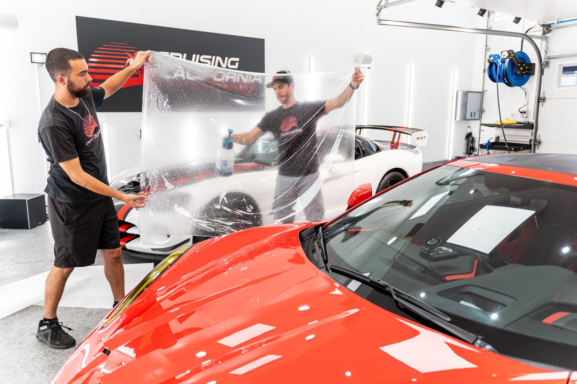Two men are wrapping a red sports car with a clear plastic sheet.
