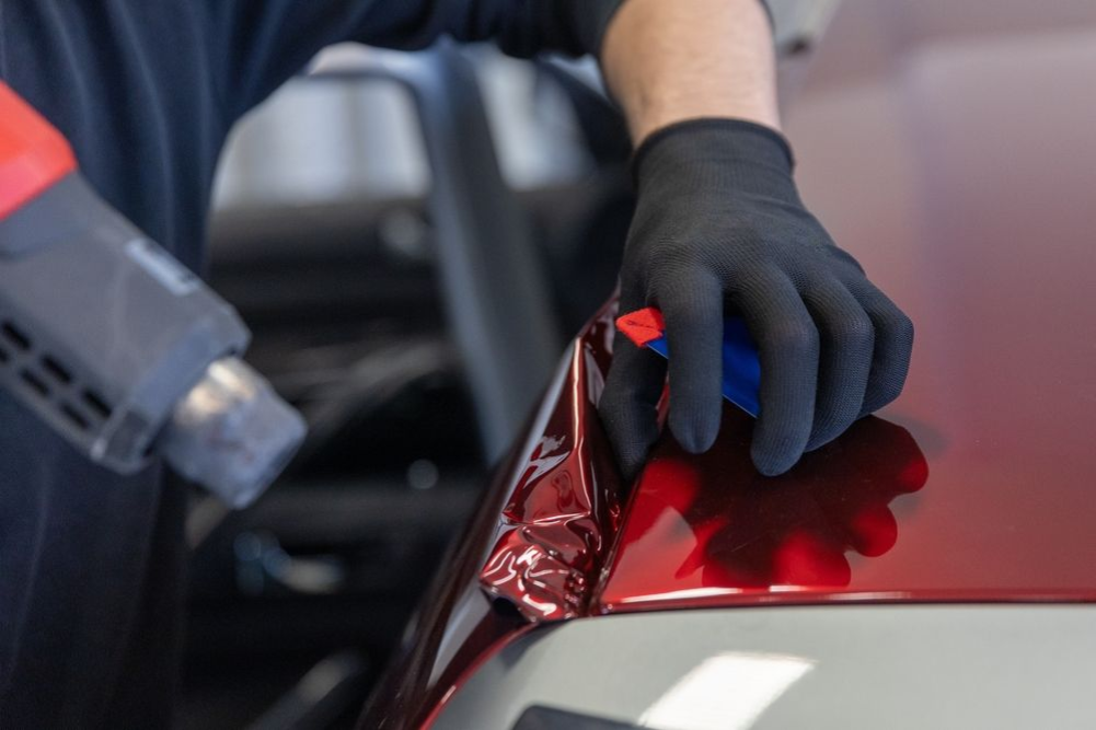 Gloved hands applying red vinyl wrap to a car panel with a heat gun