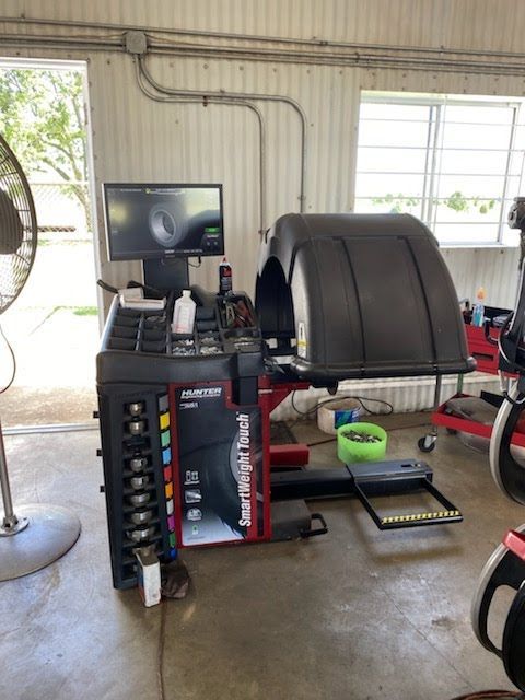 Tire balancing machine in a garage. A tire is mounted, computer screen visible, fan in the corner.