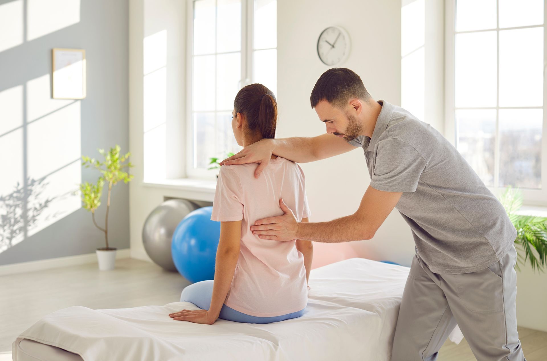 Physiotherapist examining a seated patient's back in a bright therapy room.