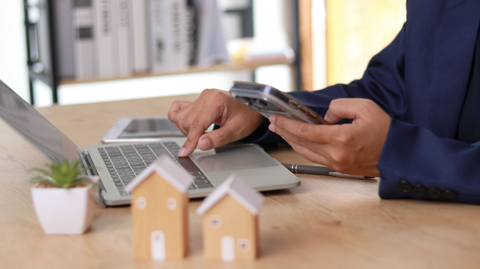 a person using a laptop and holding a smartphone at a wooden desk with two small wooden house models
