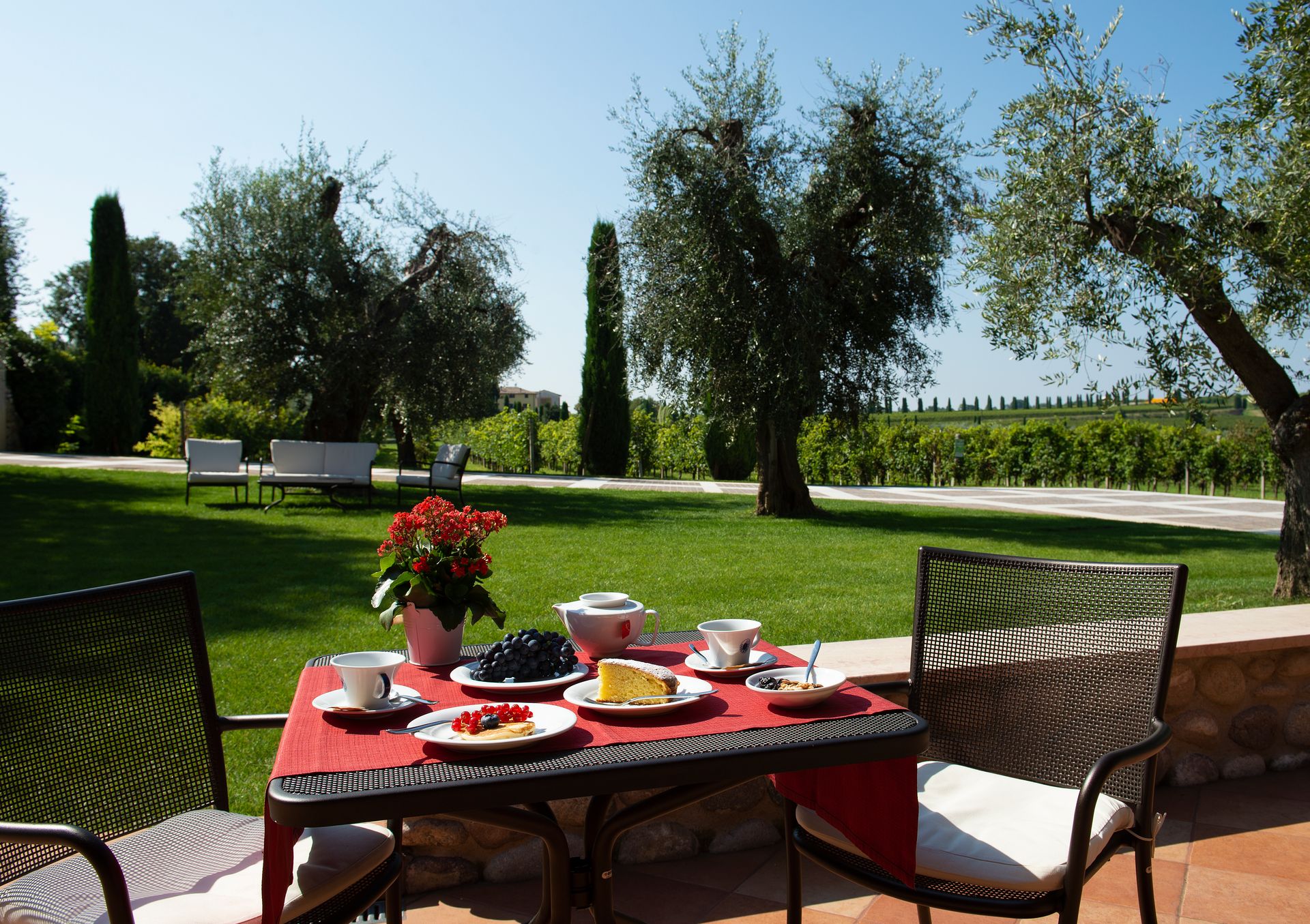 Tavolo da colazione all'aperto in patio con tovaglia rossa, fiori e sedie che si affacciano su un giardino verde.