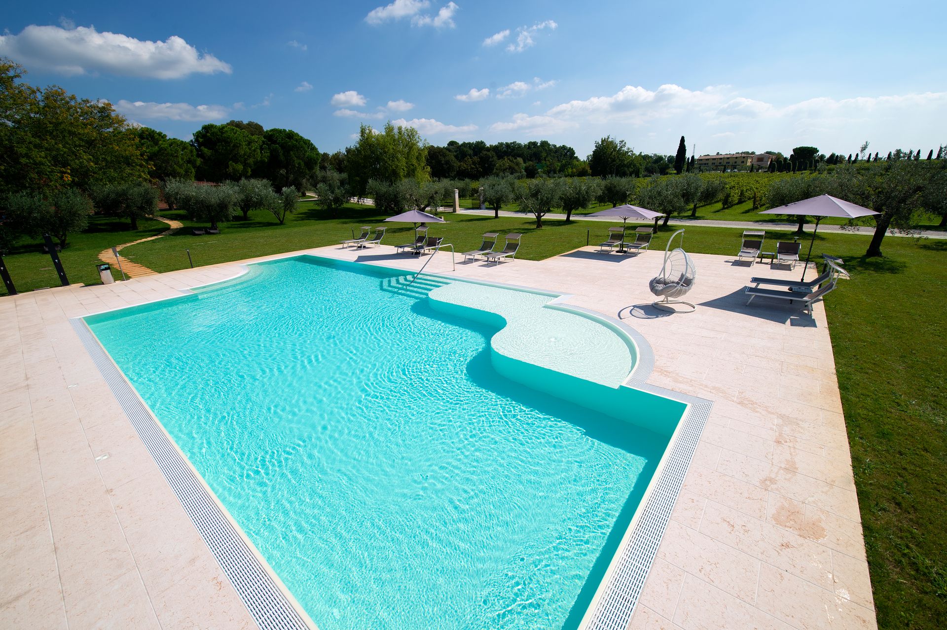 Piscina all'aperto con acqua turchese, lettini bianchi e ombrelloni in un giardino soleggiato.