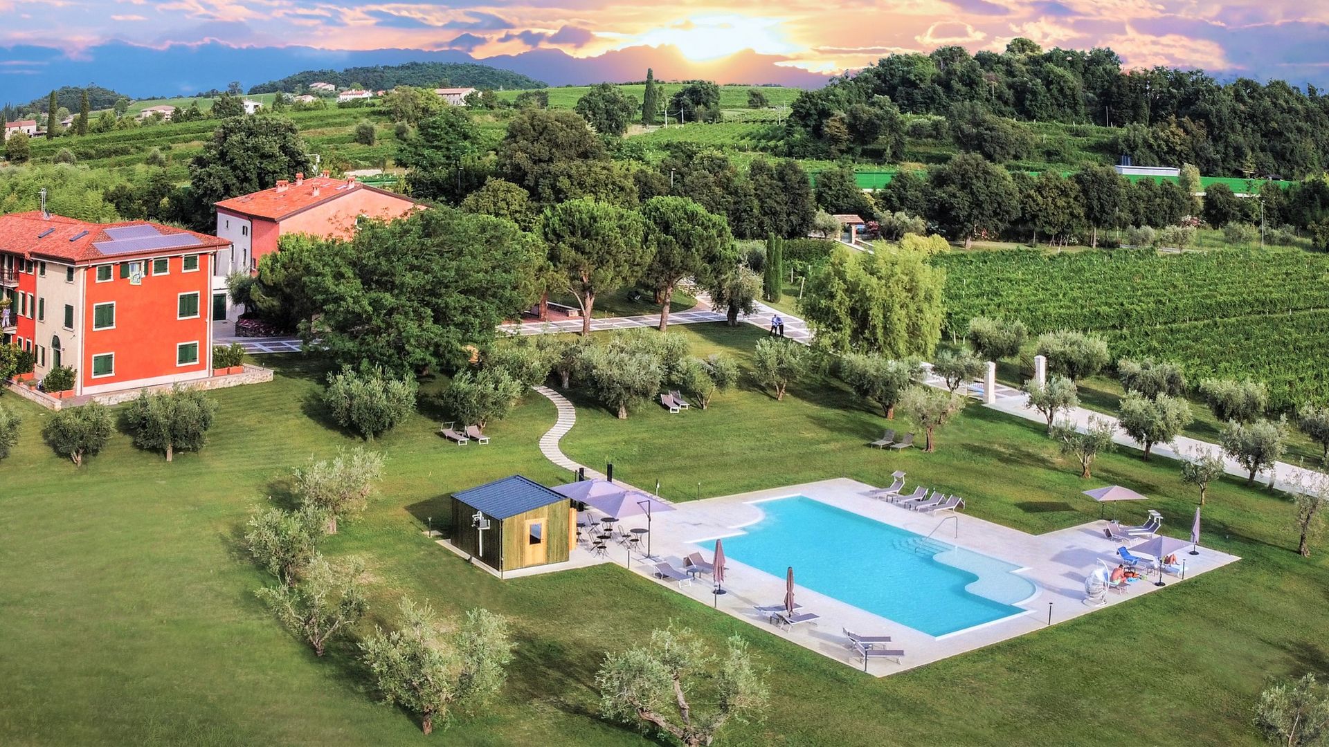 Piscina del resort accanto a prati verdi e ville al tramonto, con le colline sullo sfondo.