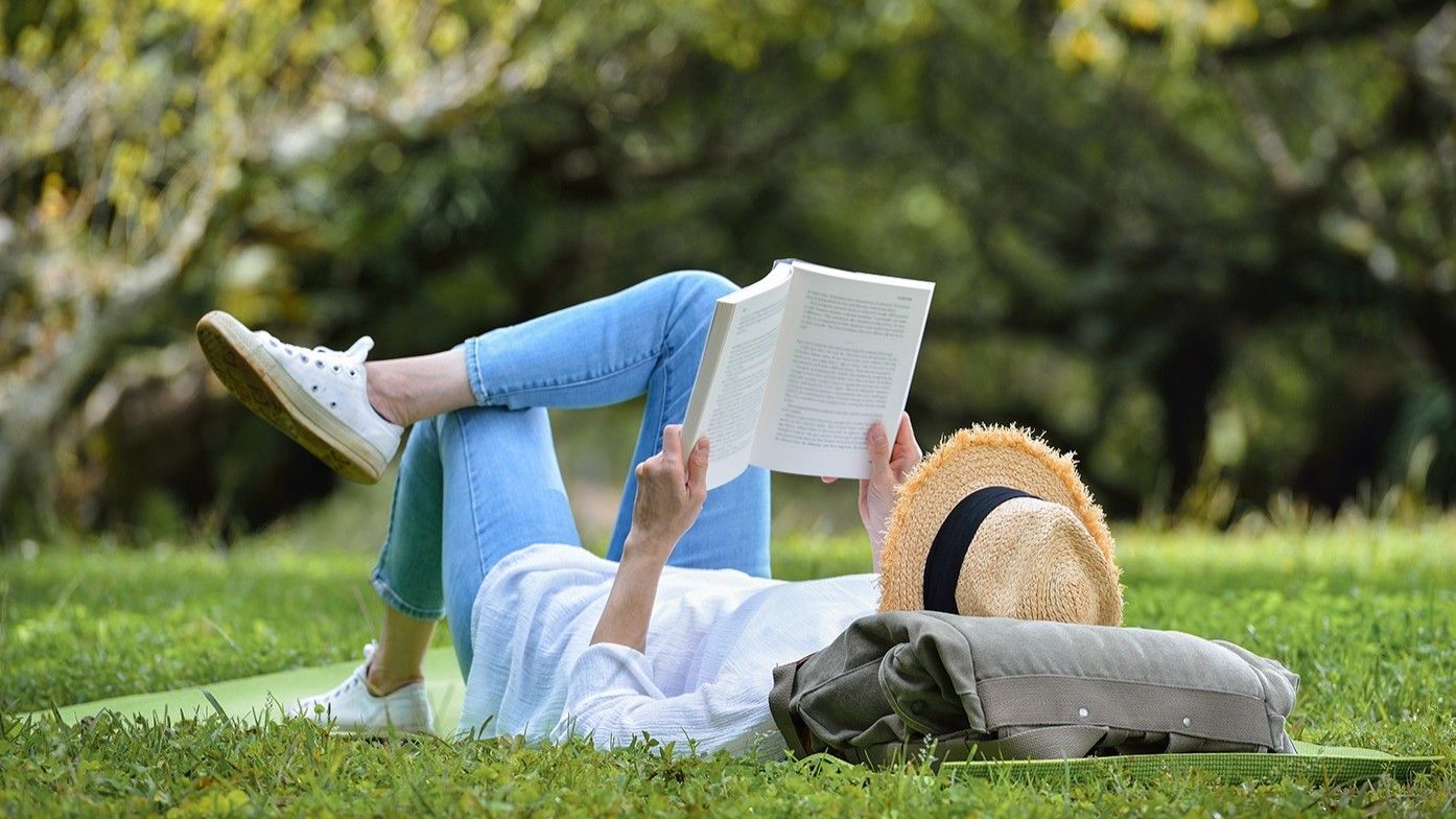 Una persona sdraiata sull'erba legge un libro in un parco soleggiato, con un cappello e uno zaino nelle vicinanze.