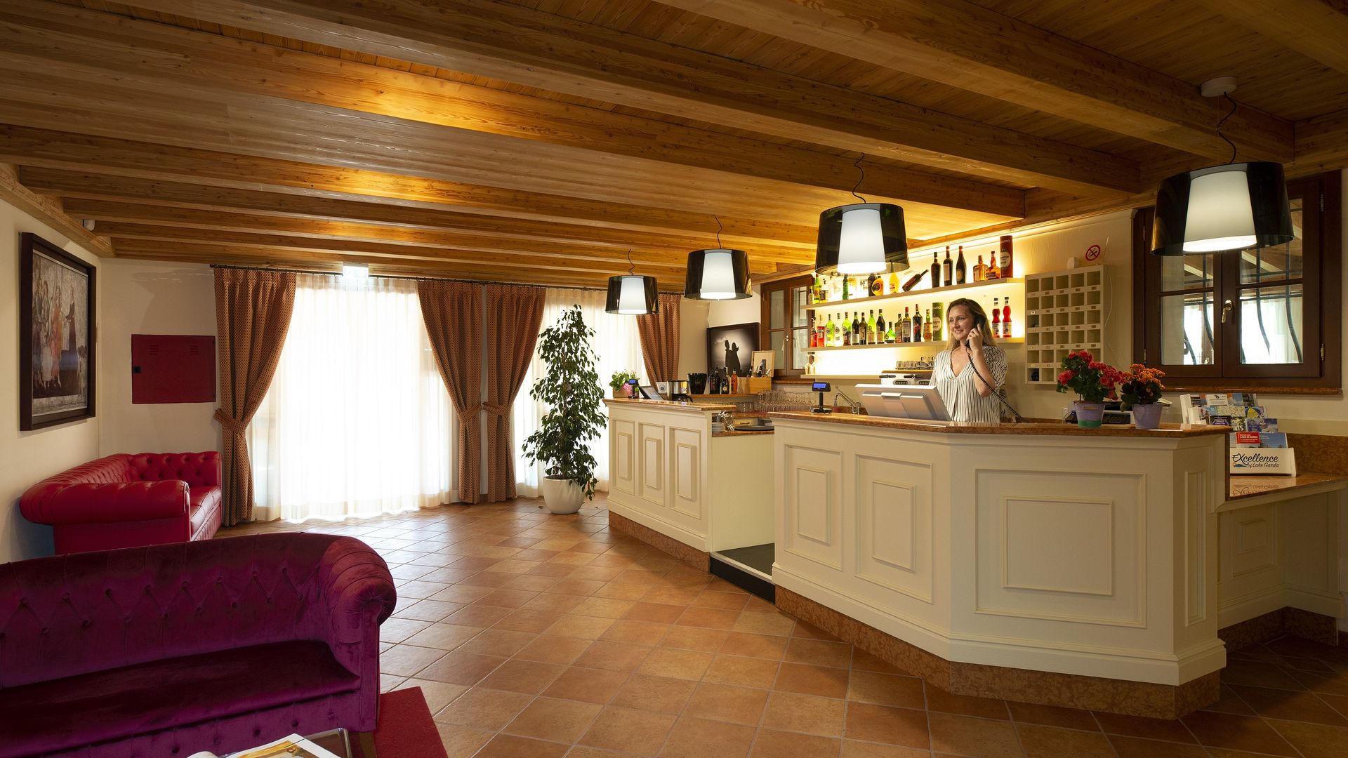 Cucina accogliente con soffitto in legno, mobili bianchi, bancone bar e divano bordeaux accanto a una finestra luminosa.