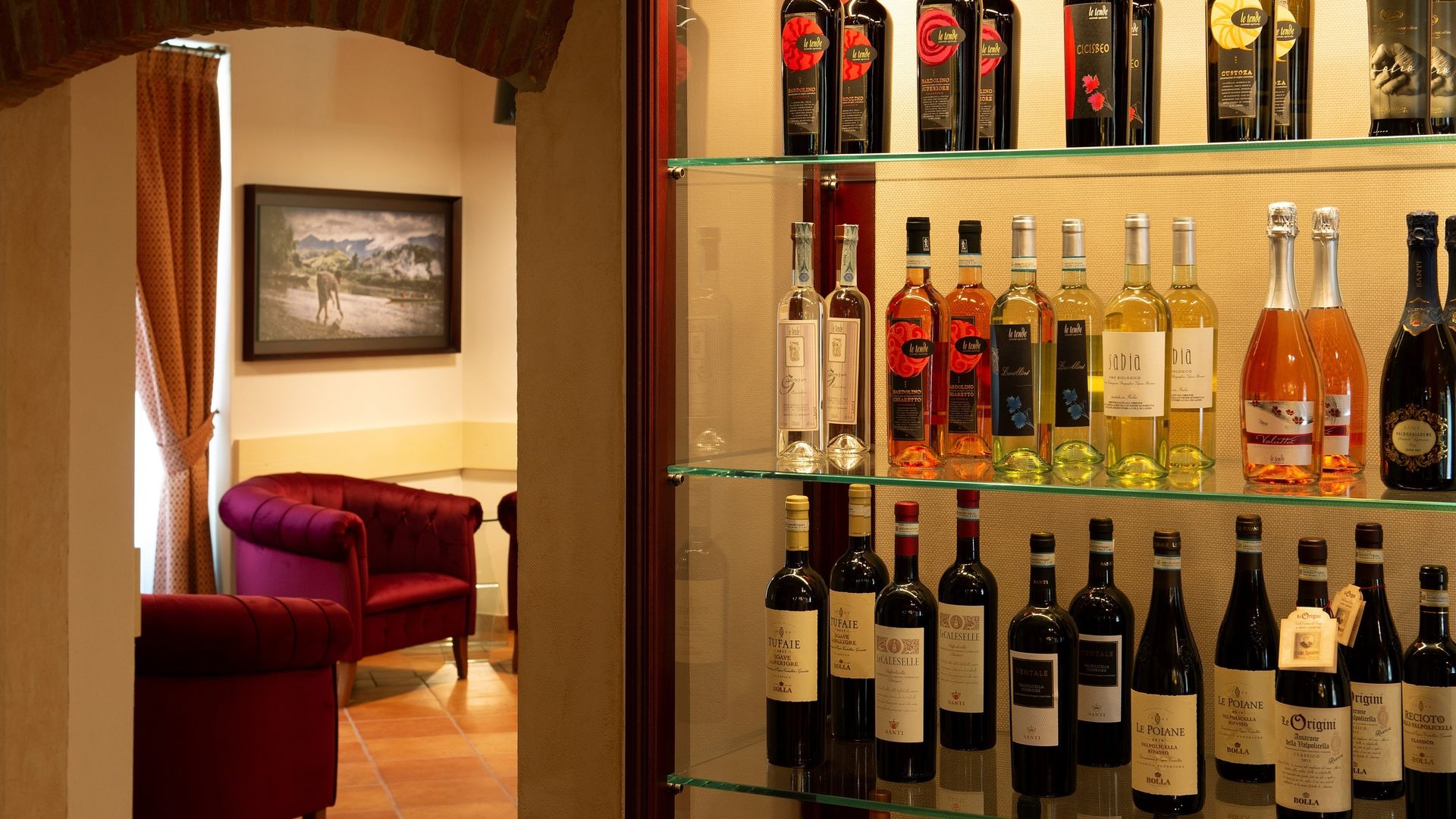 Esposizione di vini in un salotto illuminato da una luce calda, con scaffali pieni di bottiglie e una poltrona in velluto rosso.