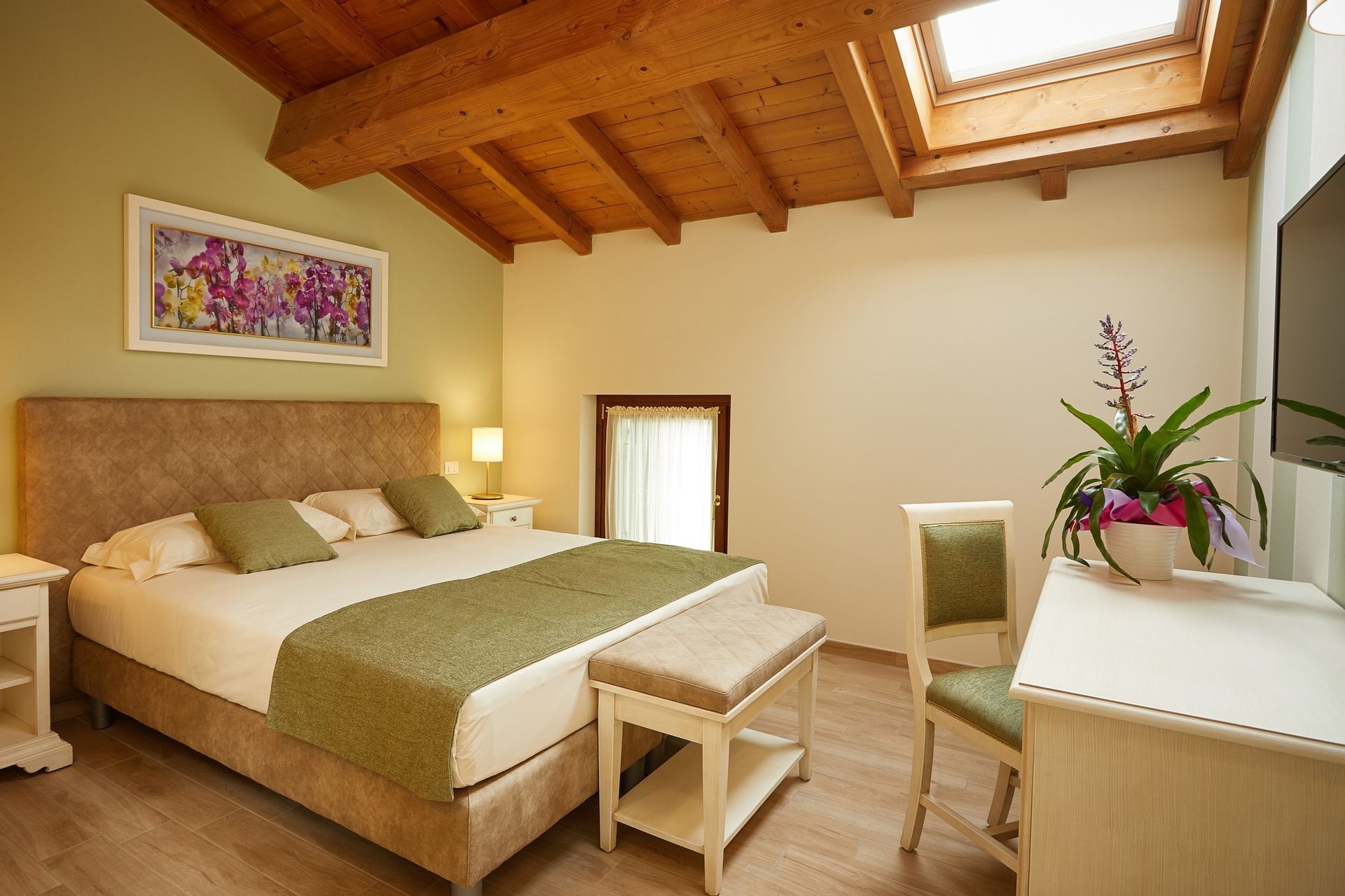 Camera da letto luminosa con soffitto con travi a vista, letto king size, panca e comò con fiori.