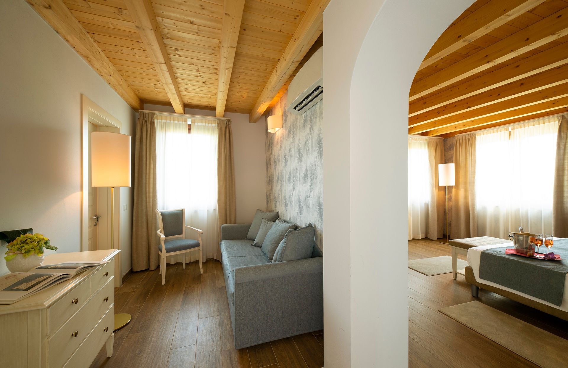Soggiorno luminoso e accogliente con divano grigio, soffitto in legno e ampie finestre che lasciano entrare la luce del sole.