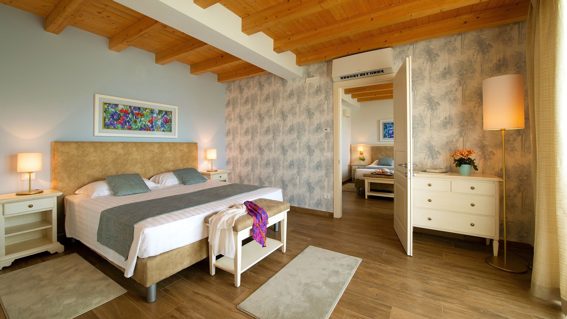 Accogliente camera da letto con letto bianco, travi in ​​legno, carta da parati a motivi geometrici e comò con lampada.