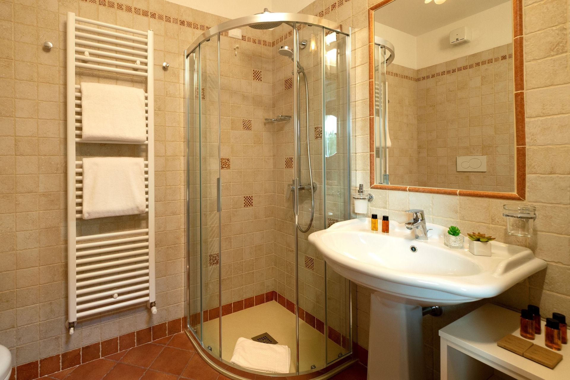 Piccolo bagno piastrellato con doccia in vetro, lavabo bianco, termoarredo e specchio con cornice in legno.