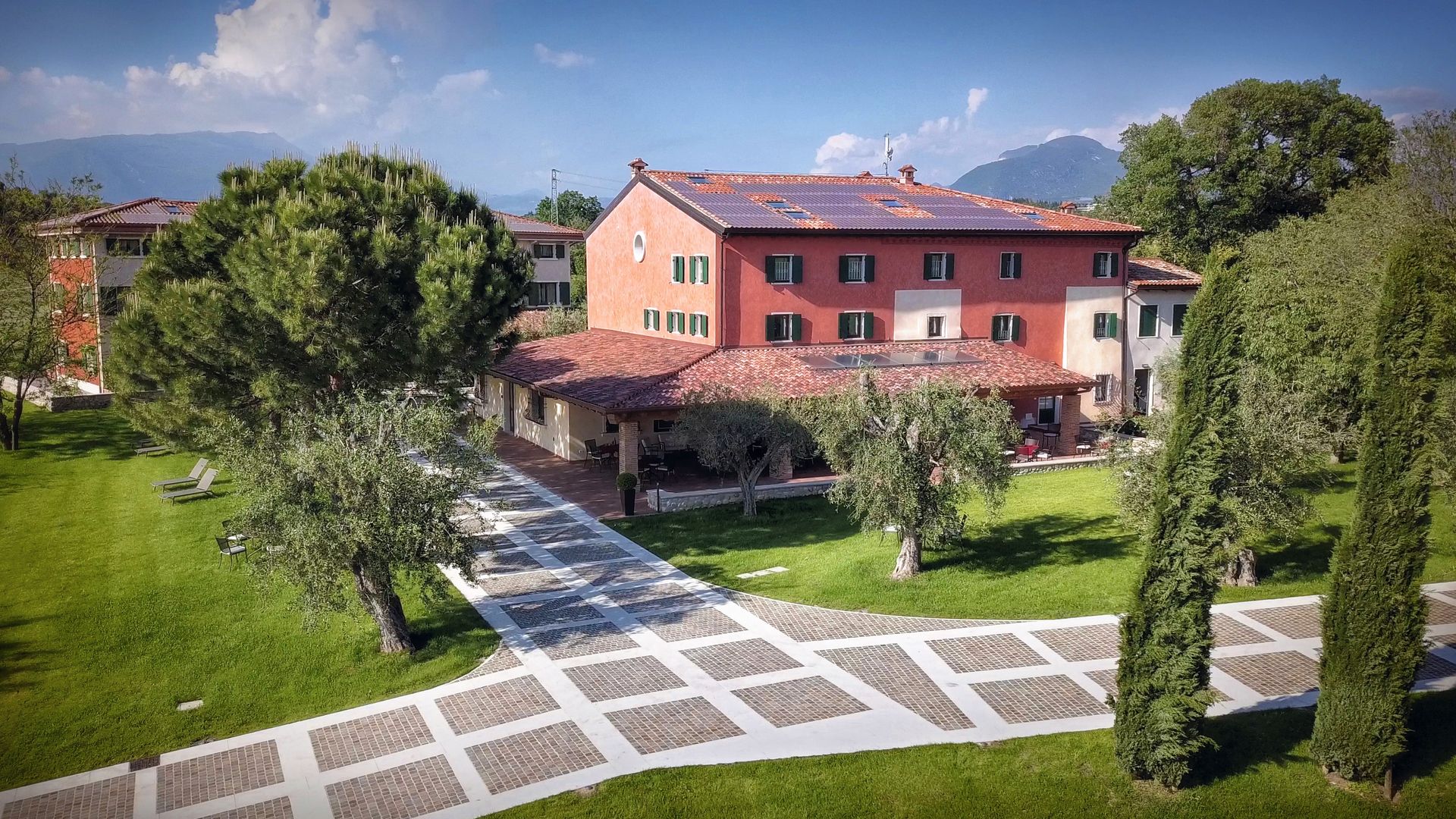 Villa rosa baciata dal sole con tetto rosso, accanto a un sentiero nel giardino e rigogliosi alberi verdi sotto un cielo azzurro.