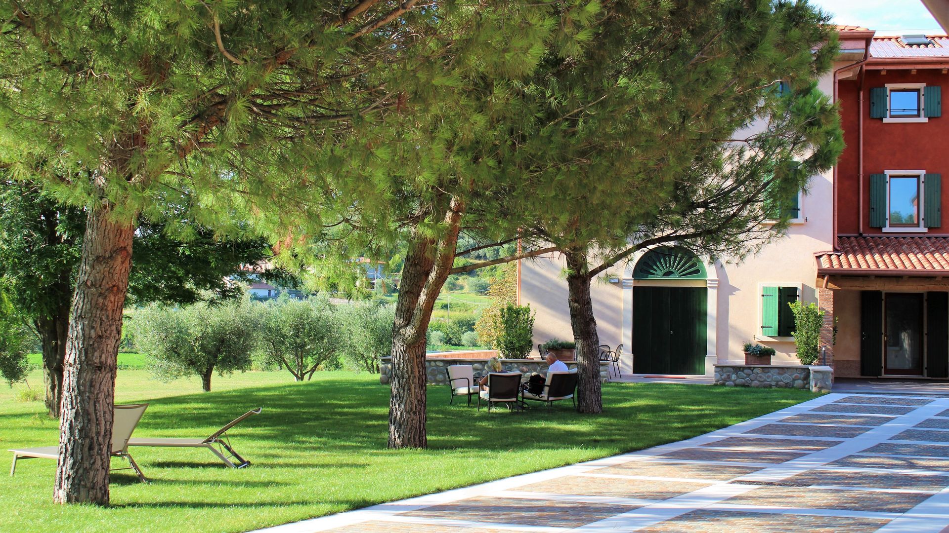 Cortile alberato con prato verde, panchine e un edificio con balconi e ingresso ad arco sulla destra.