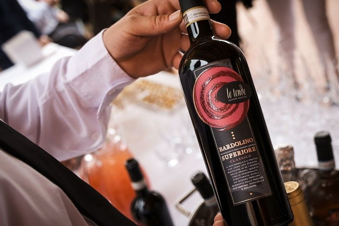 Mano che regge una bottiglia di vino nera con etichetta rosa durante una degustazione.