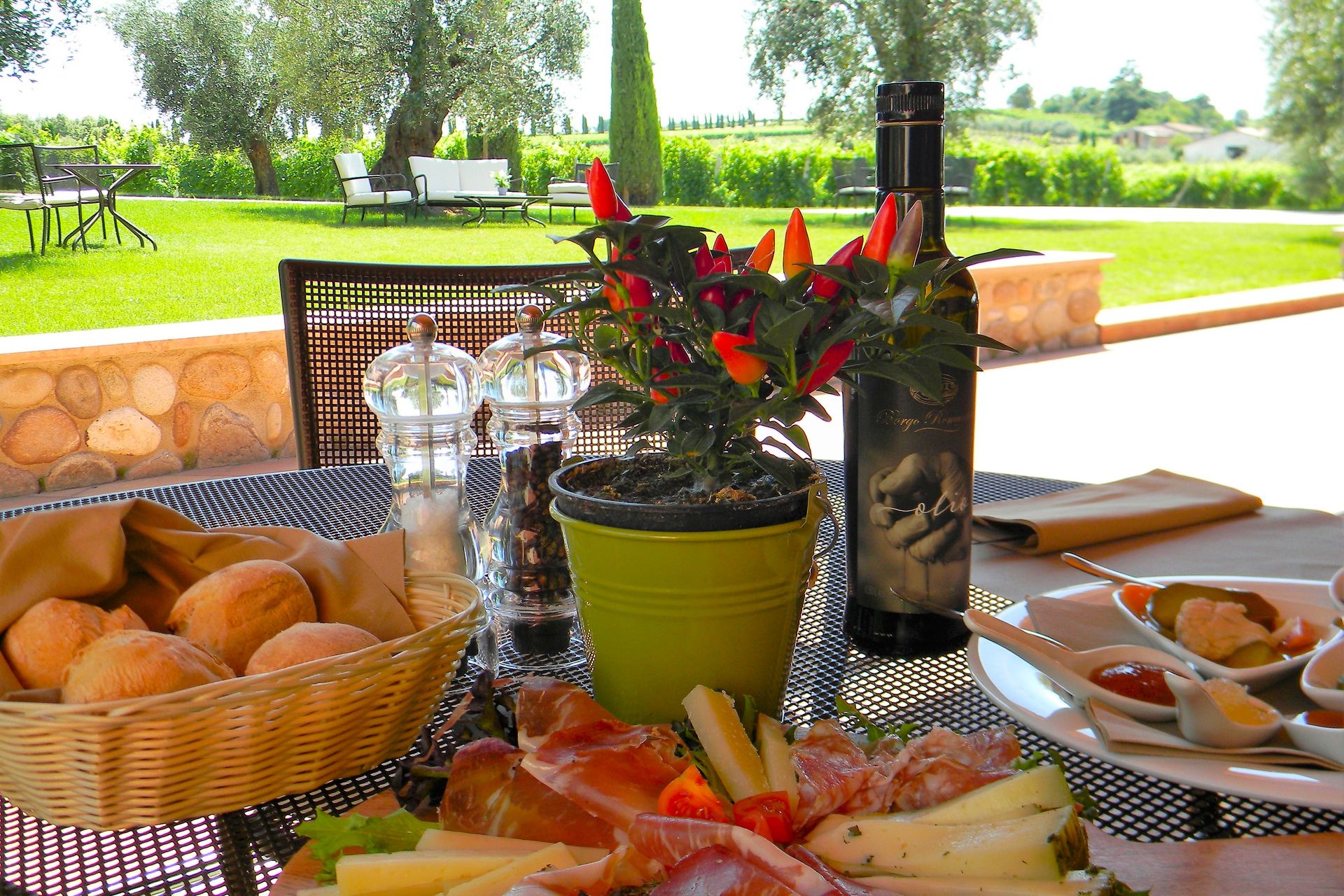 Colazione servita su un tavolo all'aperto in terrazza con pane, frutta, fiori e una bottiglia di vino, con vista su un prato verde.