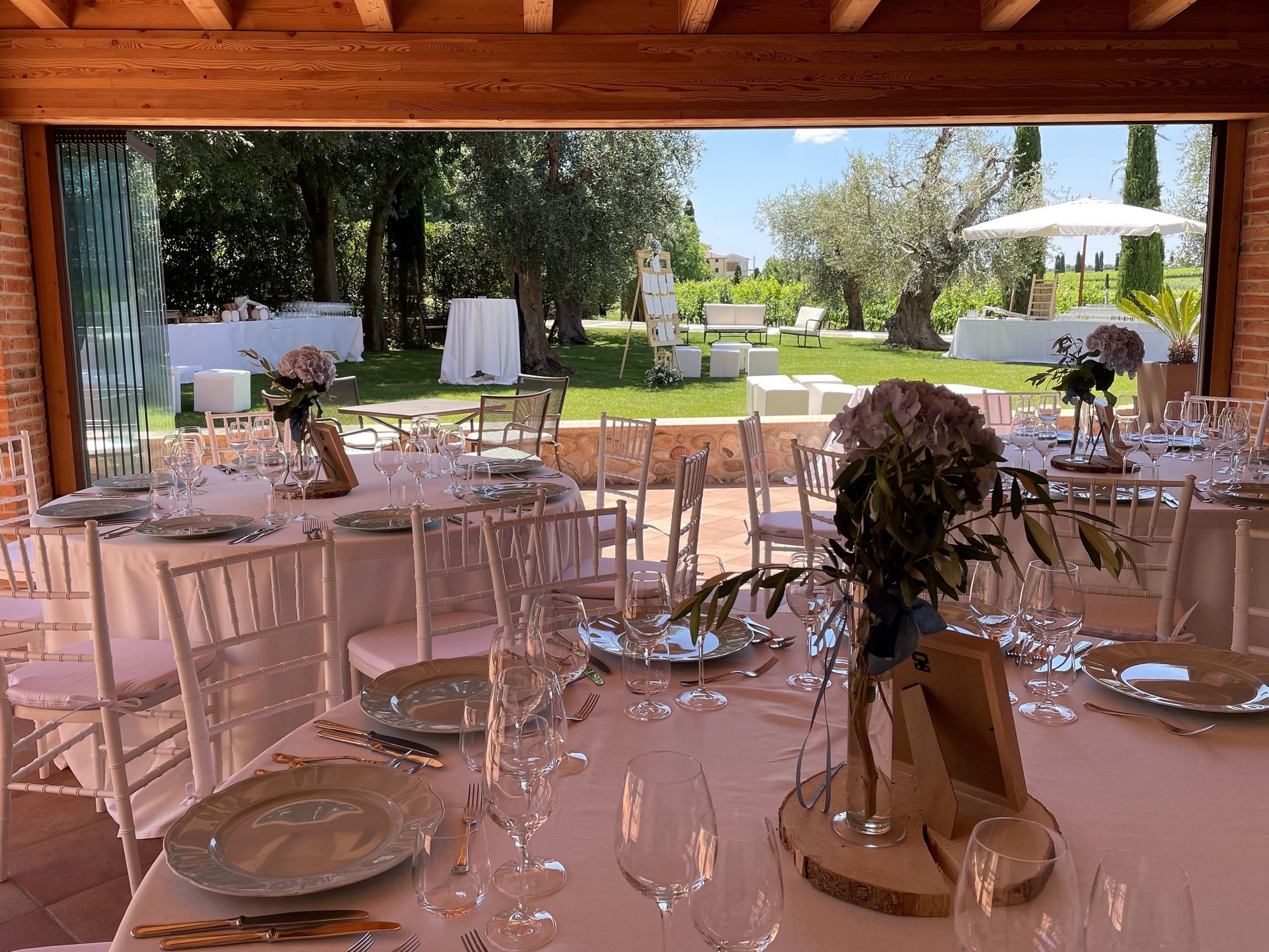 Eleganti tavoli da banchetto all'aperto, disposti sotto una tettoia di legno in un giardino soleggiato, con sedie bianche.
