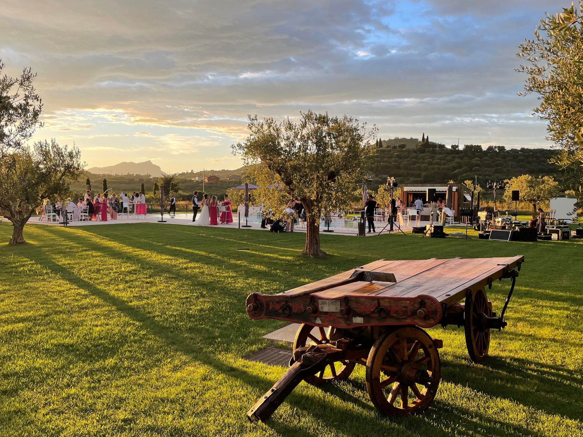 Carro rustico in un campo erboso al tramonto, con un gruppo di persone e degli alberi sullo sfondo.