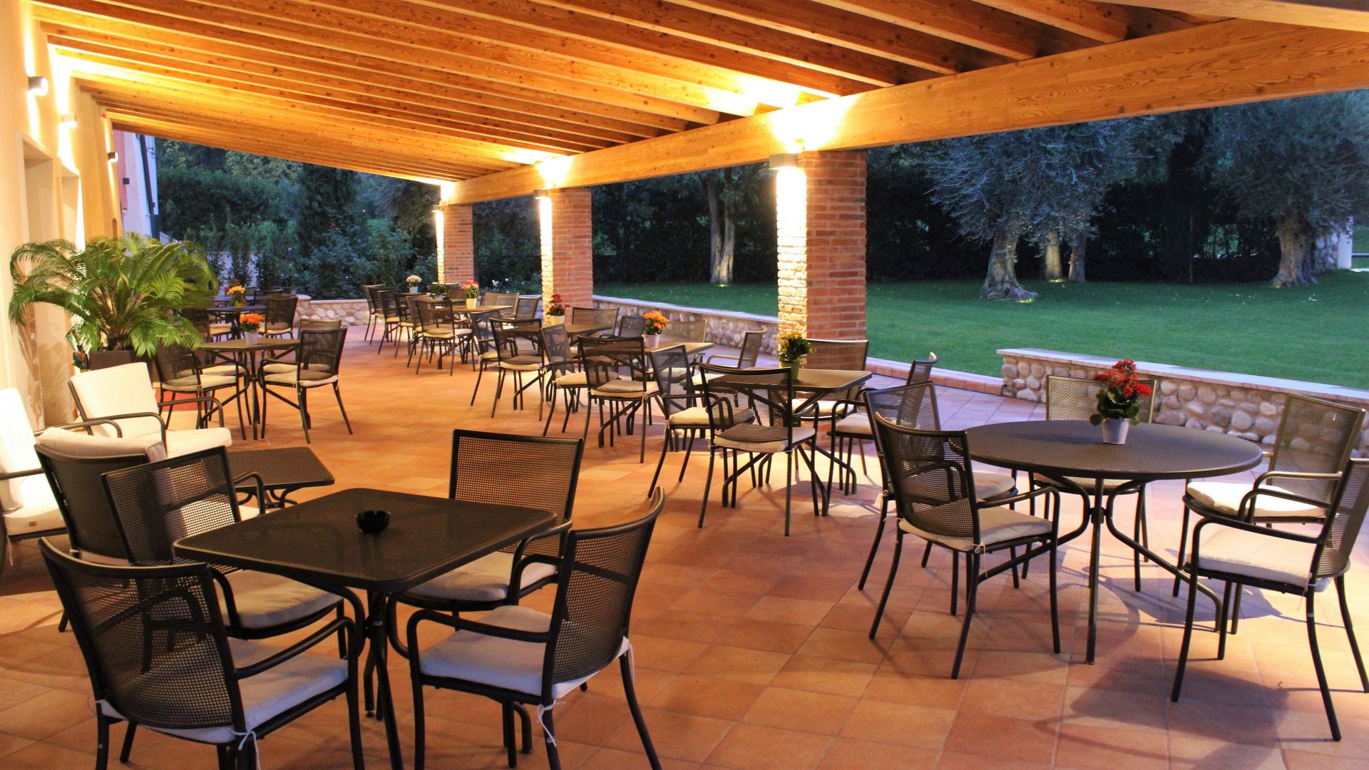 Patio coperto con tavoli e sedie in vimini, luci soffuse e vista su un prato al crepuscolo.