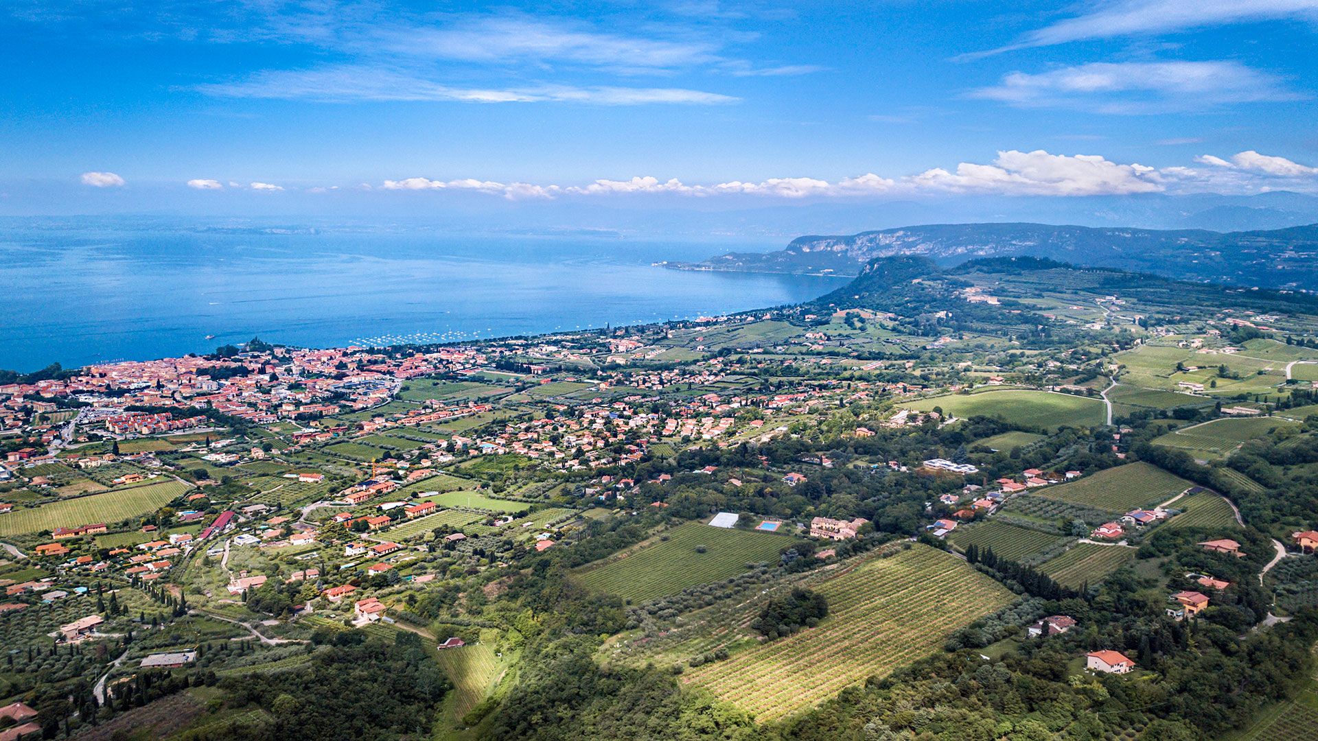Veduta aerea di una cittadina costiera con case dai tetti rossi, colline verdi e il mare blu sotto un cielo limpido.