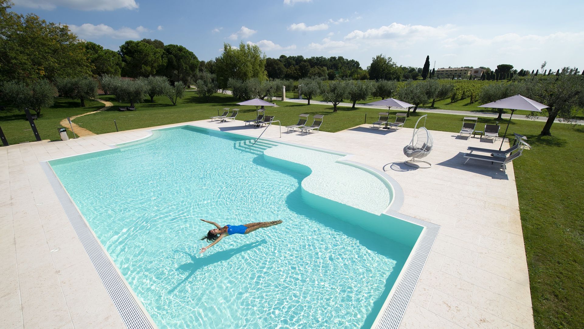 Piscina all'aperto con acqua turchese, una persona che galleggia e lettini prendisole su un prato soleggiato con ombrelloni.