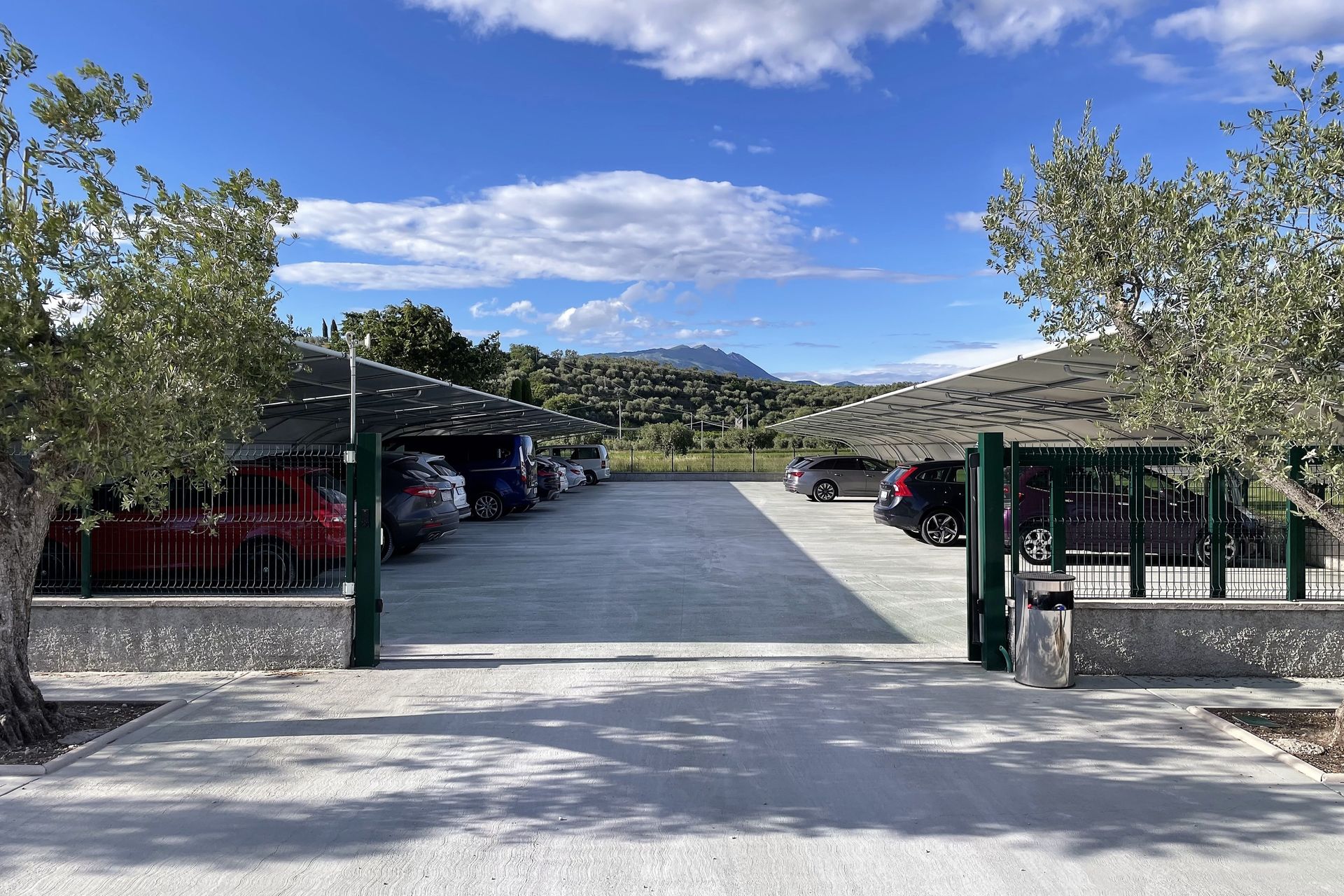 Apri il cancello che conduce a un vialetto di ghiaia fiancheggiato da auto parcheggiate, alberi e colline sotto un cielo azzurro.