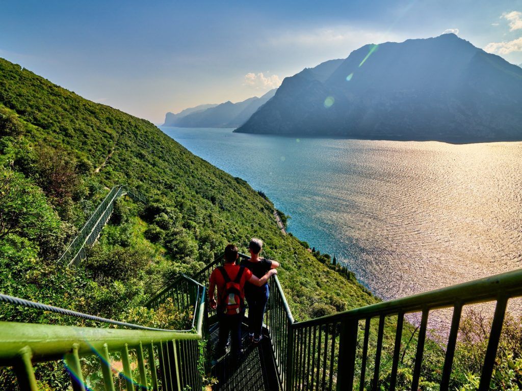 Due escursionisti scendono dei gradini verdi verso un lago illuminato dal sole, con le montagne sullo sfondo.