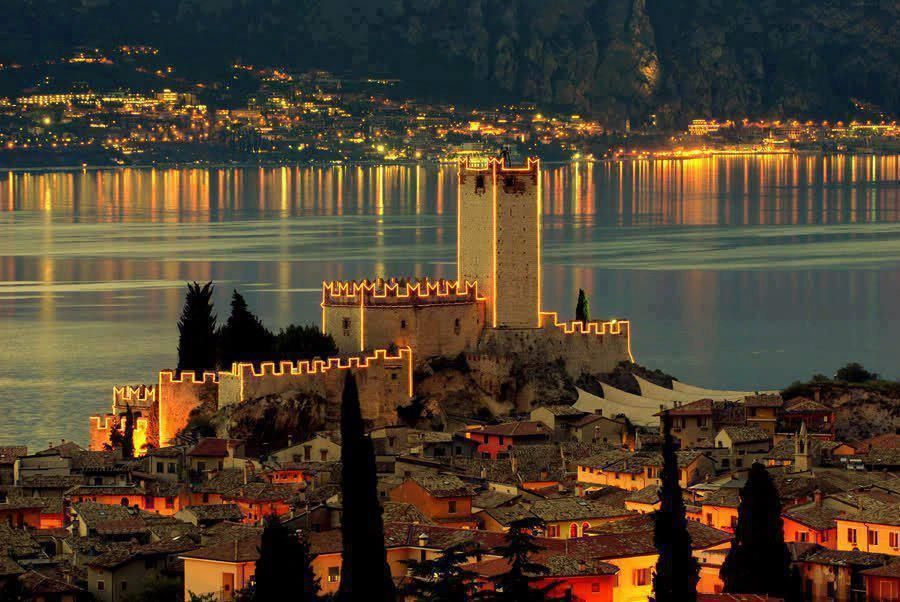 Veduta notturna di un castello e di un villaggio in riva a un lago, con luci dorate che si riflettono sull'acqua calma.