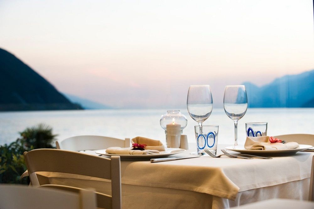 Un romantico tavolo apparecchiato in riva al lago al tramonto, con bicchieri da vino e piatti, con vista sulle acque tranquille e sulle montagne.