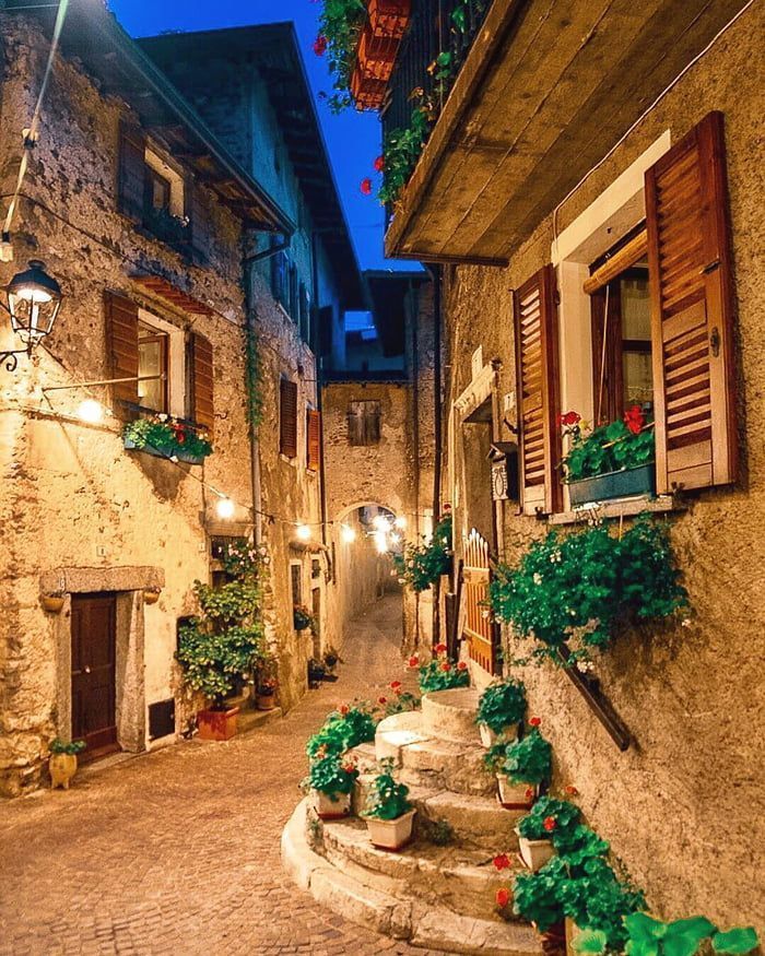 Stretto vicolo acciottolato al crepuscolo con edifici in pietra, lanterne calde e vasi di fiori sui gradini