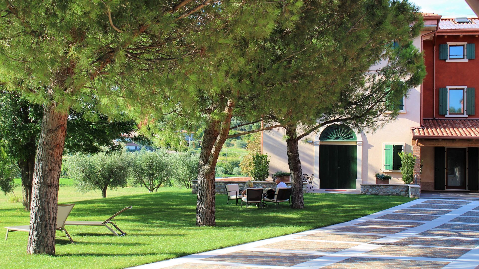 Tree-lined lawn beside a red building with benches and a paved walkway