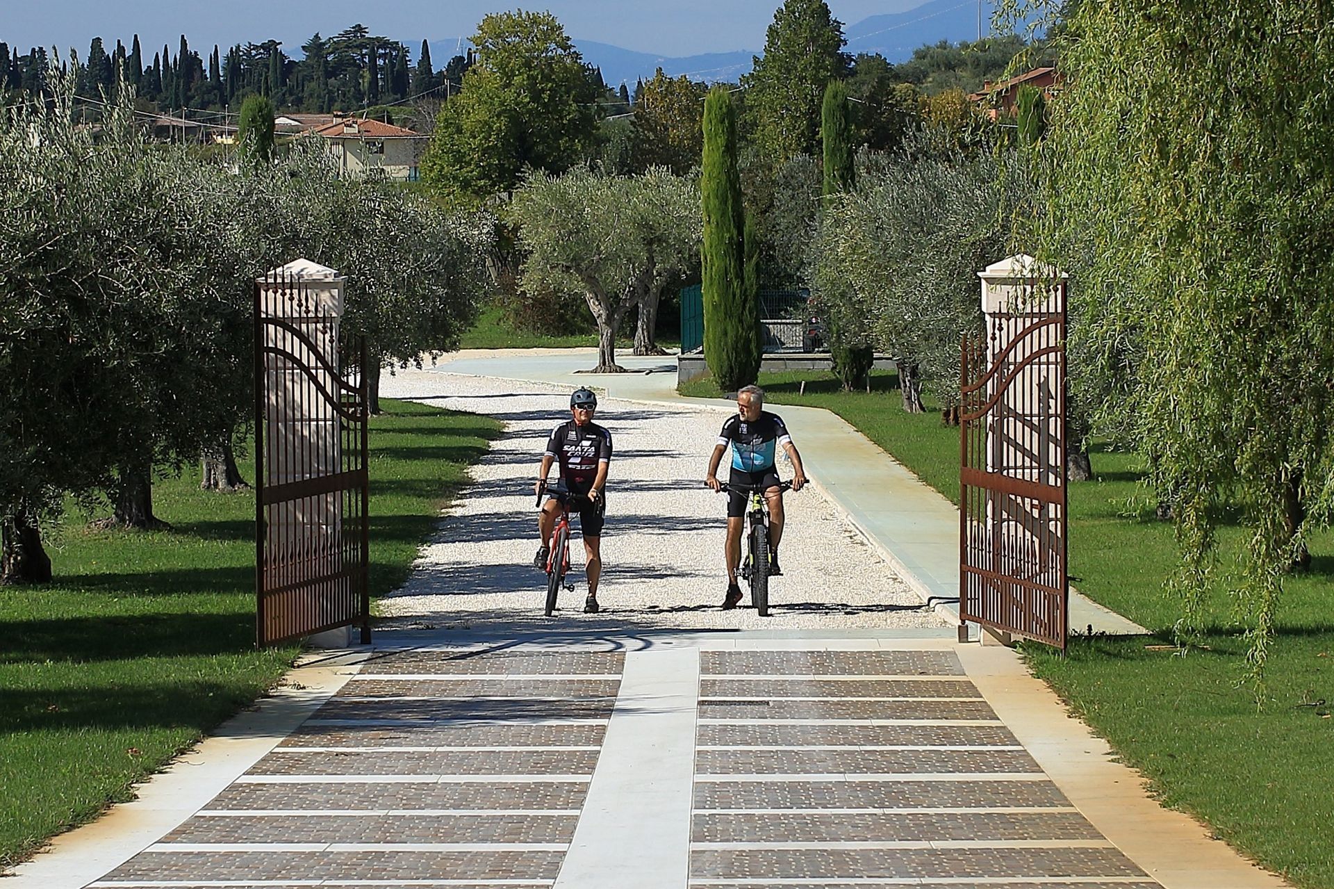 Due ciclisti percorrono un sentiero di pietra che attraversa un cancello di un giardino e un frutteto verdeggiante.